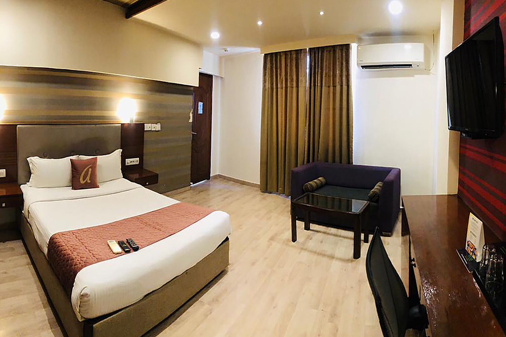 A hotel room with a large bed, a small sofa, a desk with a chair, an air conditioning unit, and a television.