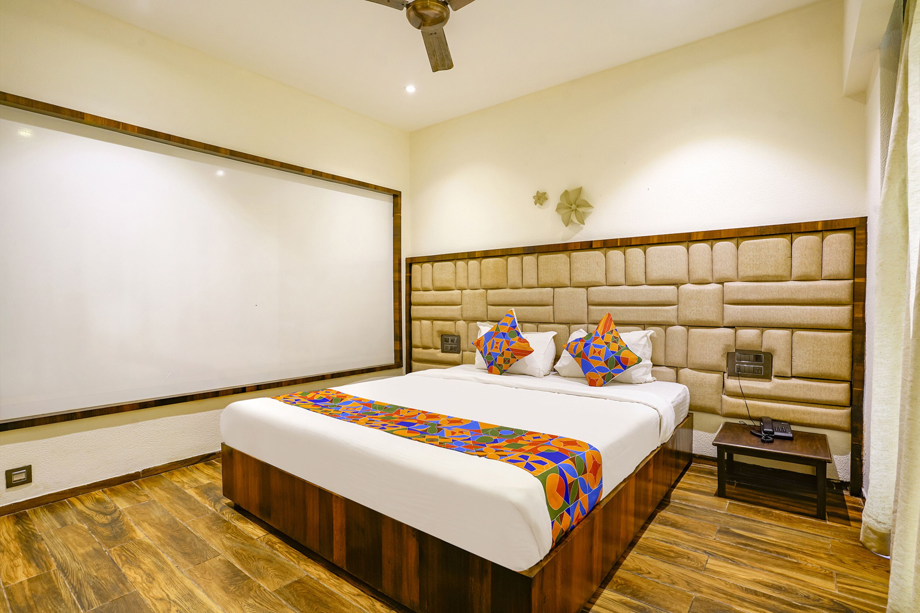A hotel room with a large bed, a headboard with a built-in TV, a bedside table, and a wooden floor.