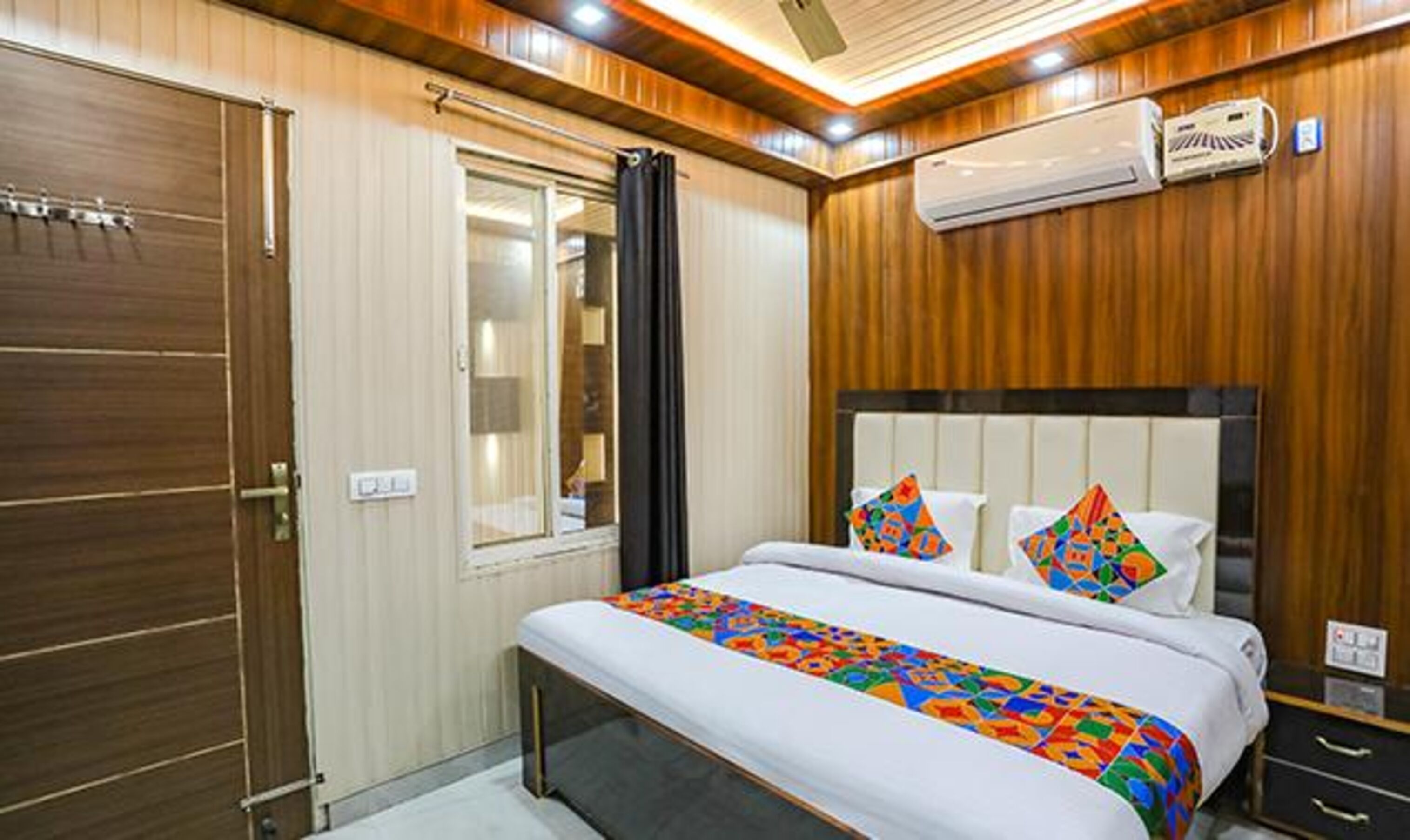 Una habitación de hotel con un revestimiento de madera, una cama con ropa de cama colorida, una ventana con cortinas y un equipo de aire acondicionado.