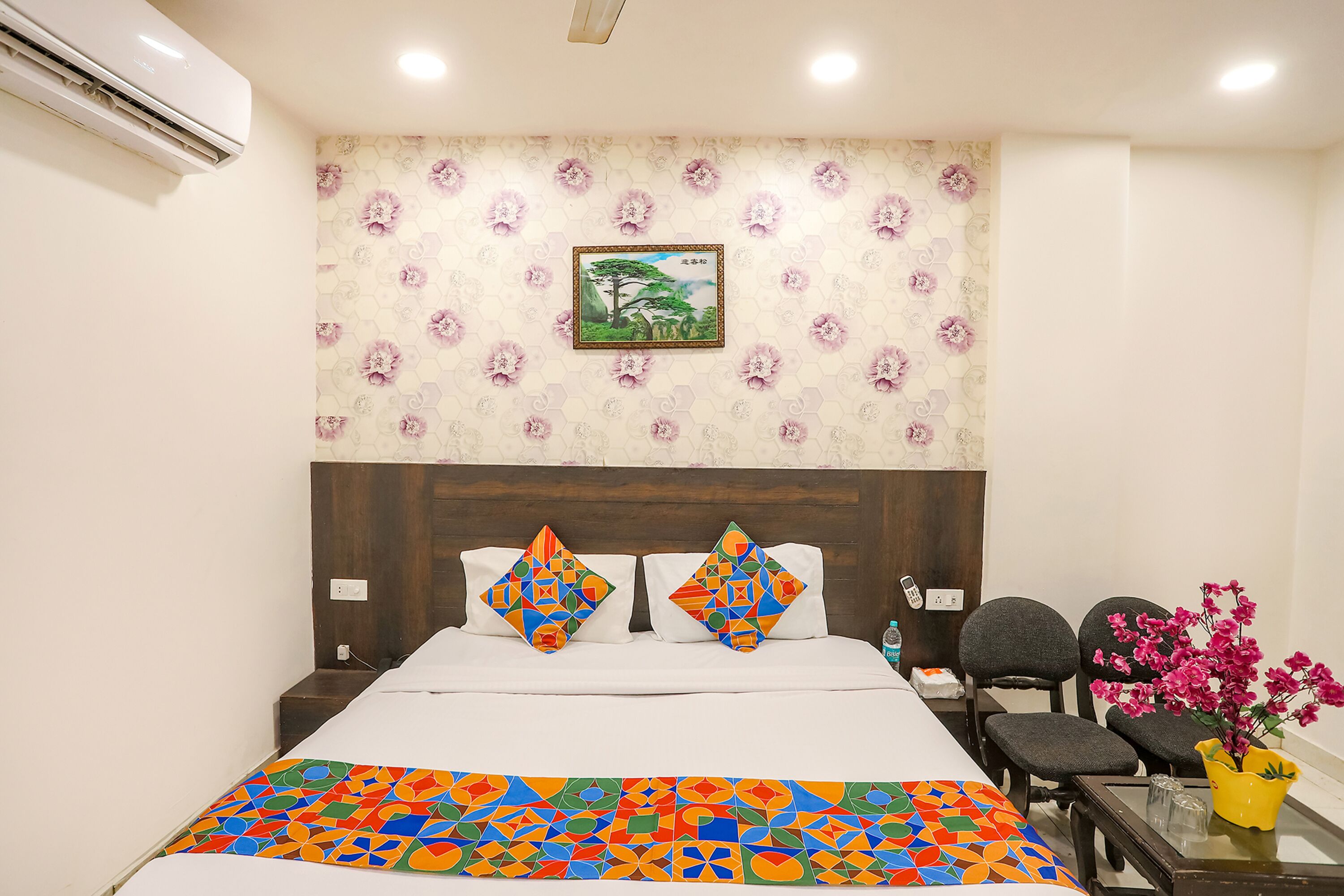 Una habitación de hotel con una cama, dos sillas, una mesita con un florero y una pared decorada con un estampado floral y un cuadro enmarcado.