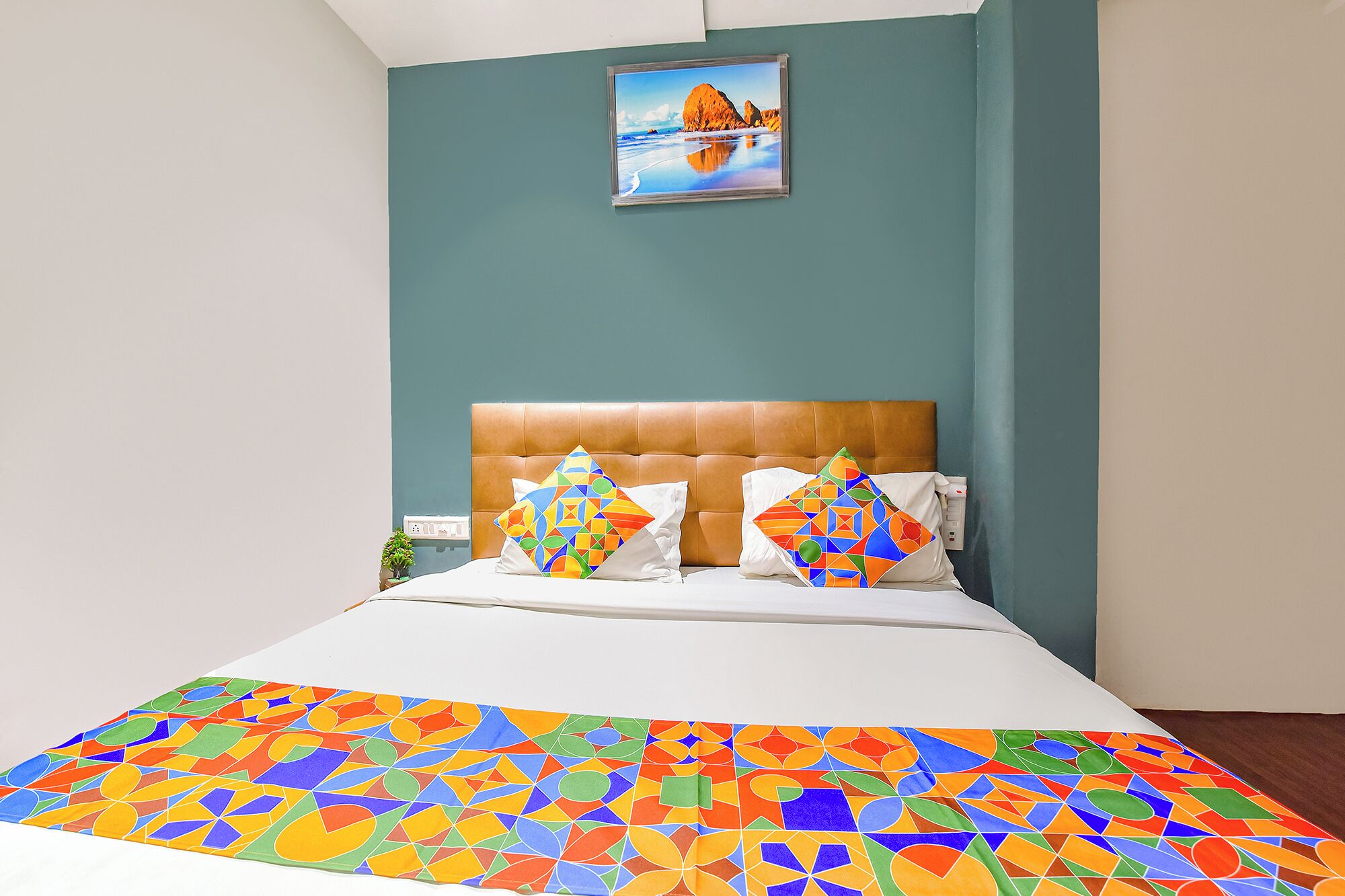 Un dormitorio con pared color turquesa, un cuadro enmarcado, cabecero marrón, dos almohadas con diseños geométricos y una colcha colorida.