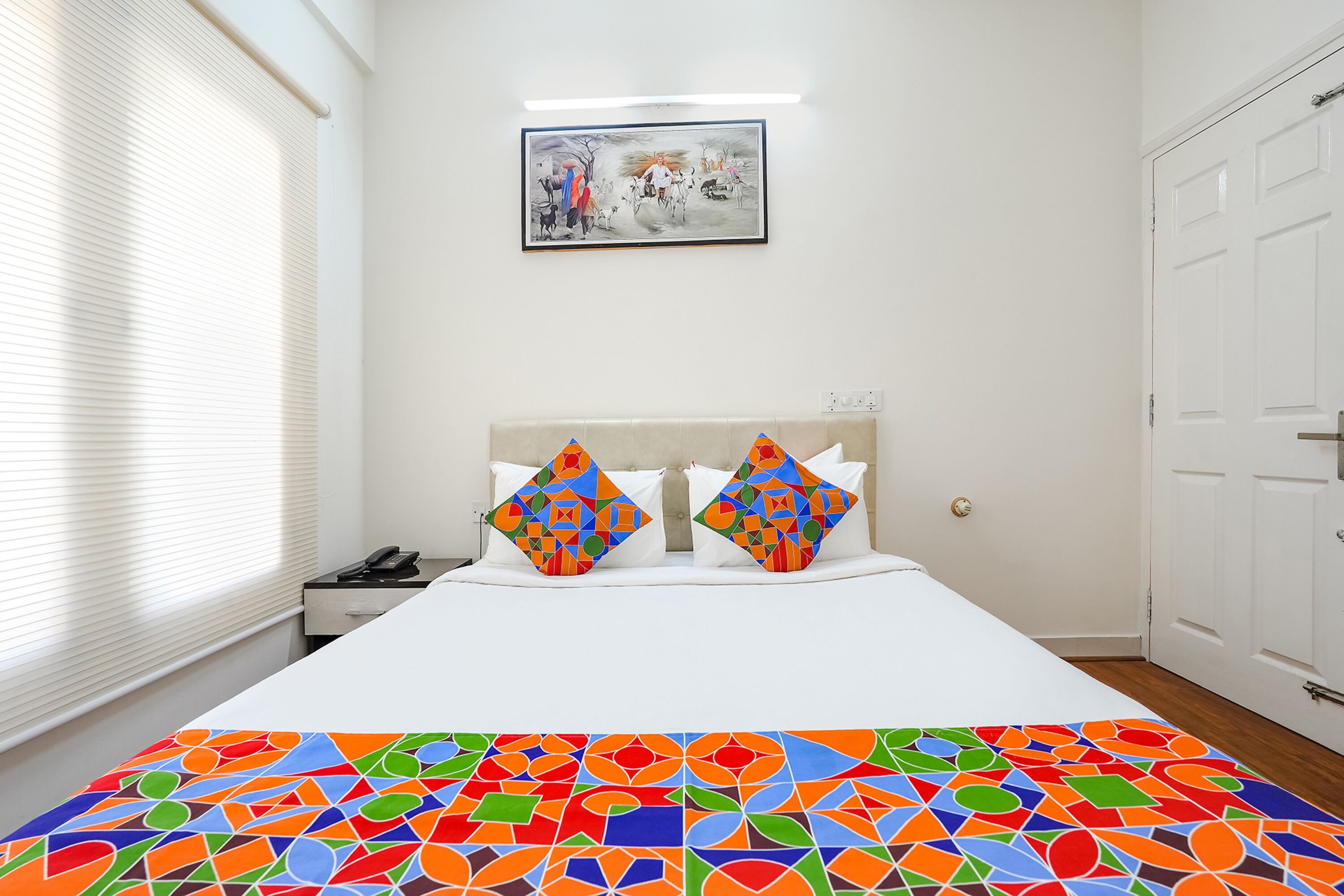 A hotel room with a bed, two colorful pillows, a painting on the wall, and a window with blinds.