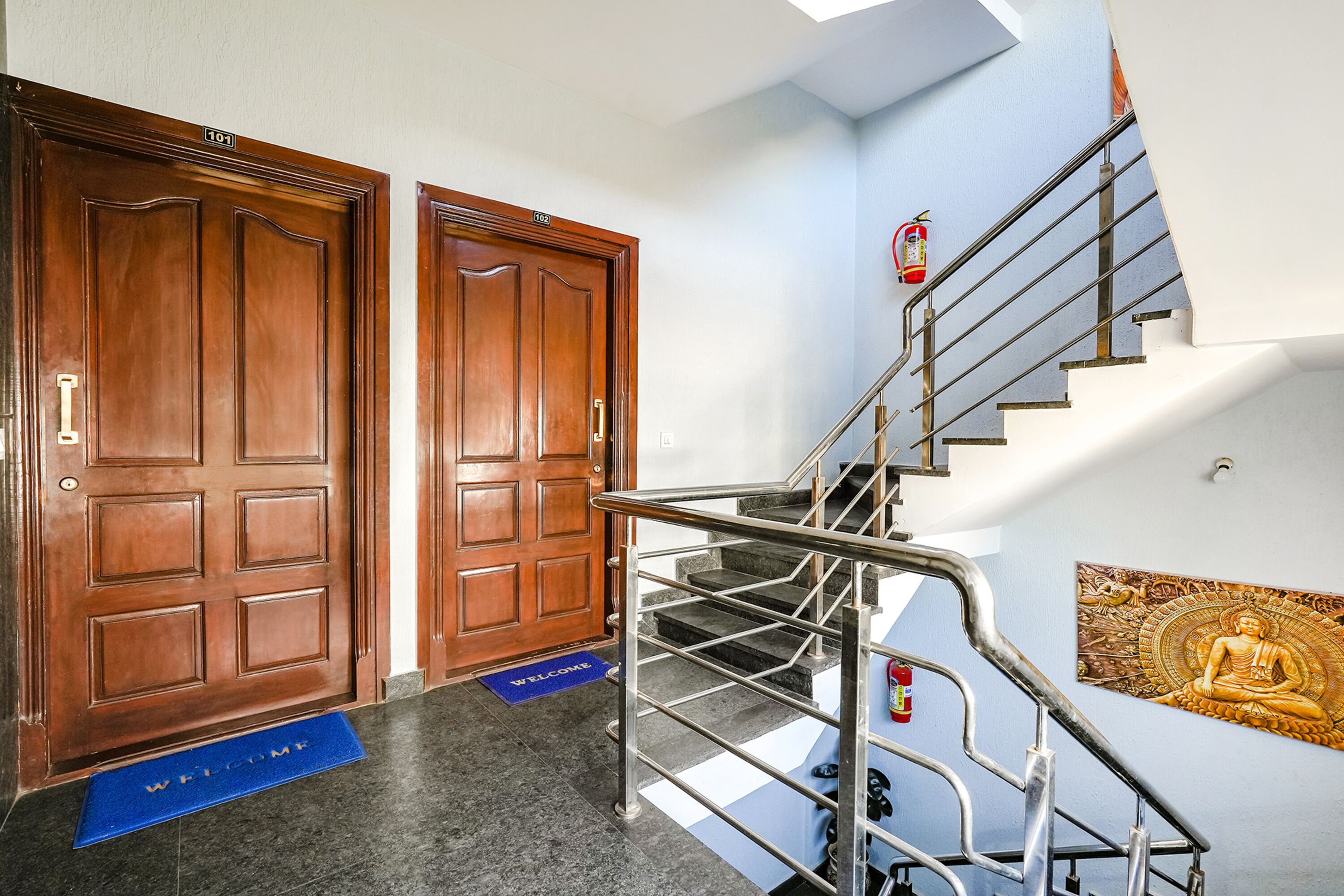 A staircase with metal railings, two wooden doors with handles, a welcome mat, and a fire extinguisher on the wall.