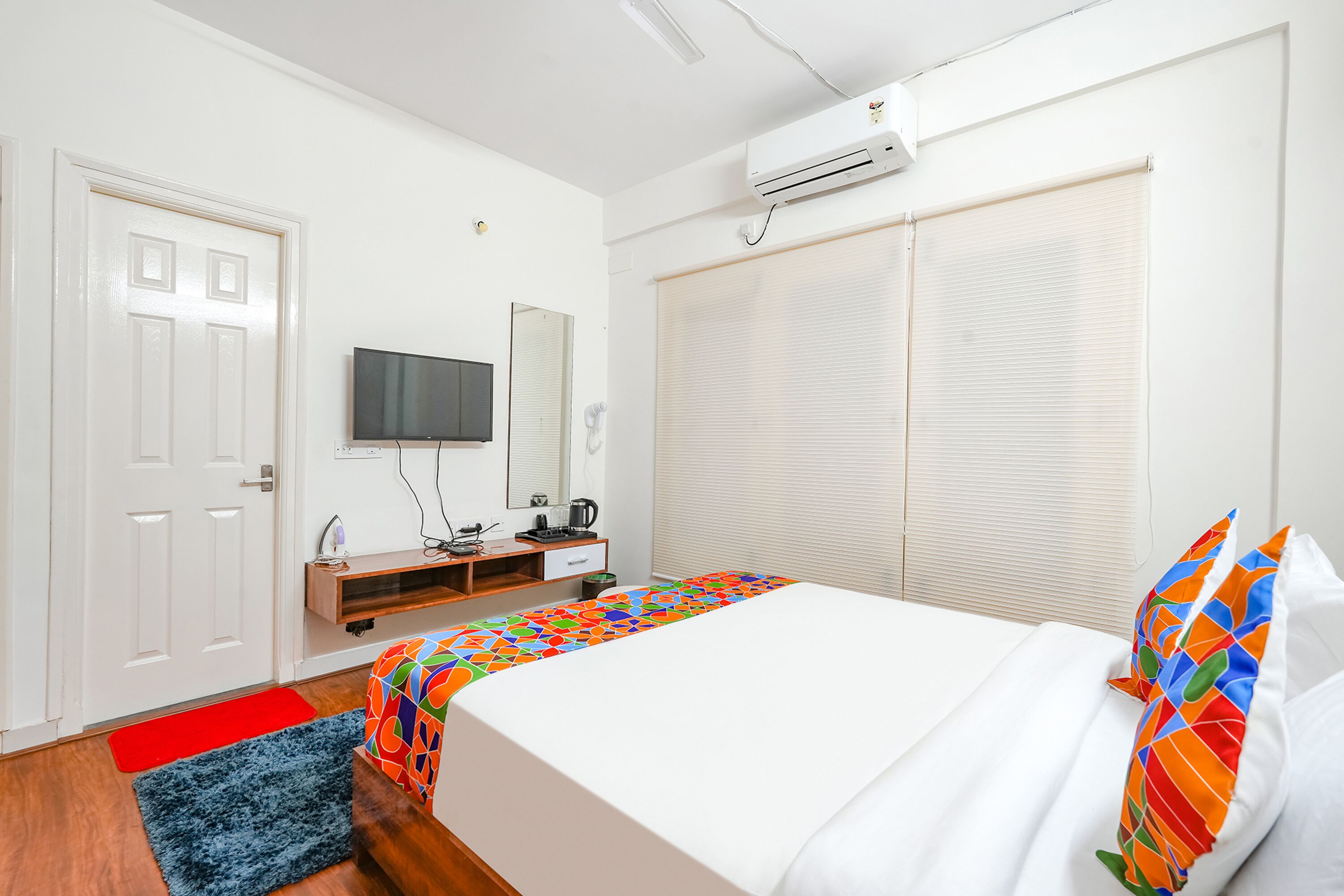 A bedroom with a bed, a TV, a desk, and a window with blinds.