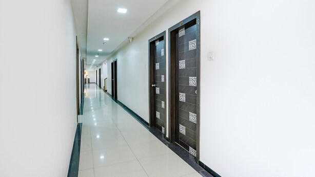 A long, narrow hallway with white walls and a tiled floor. There are doors with dark frames and decorative patterns on the doors. - Fabexpress Rajmahal (Navi Mumbai)