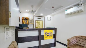 A reception desk with a sign for 'fab hotels', a ceiling fan, an air conditioning unit, and a reception area with a sofa and a chair. - Fabexpress Rajmahal (Navi Mumbai)