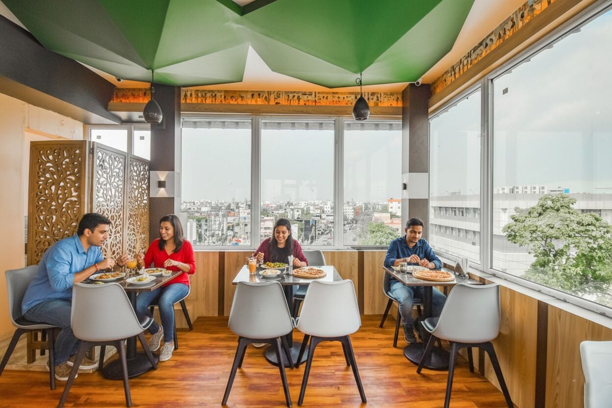 Un comedor con cuatro personas sentadas en mesas, ventanas amplias y vista a la ciudad.
