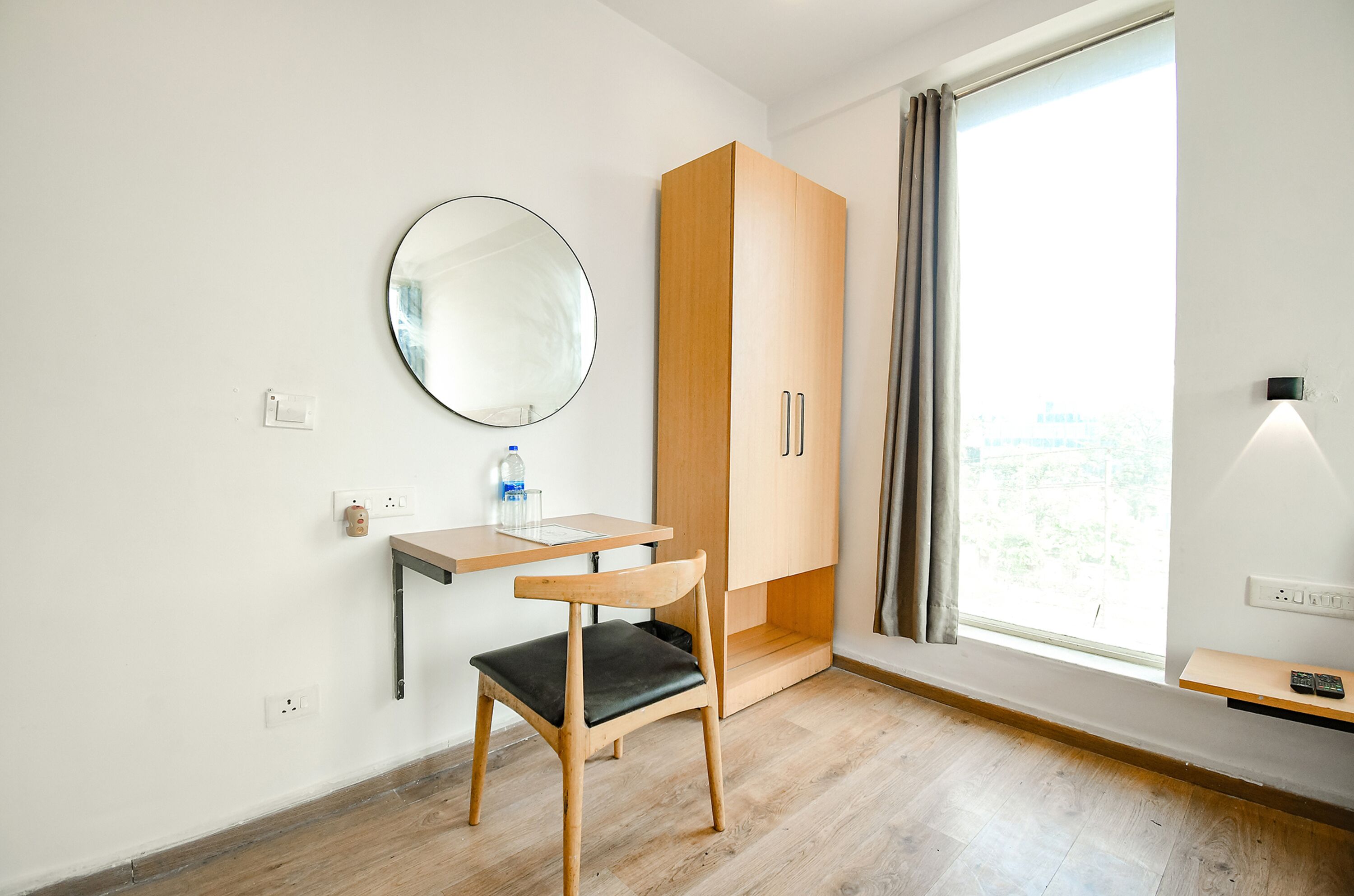 A small, modern room with a wooden desk, a chair, a wardrobe, a round mirror, a window with curtains, and a bedside lamp.