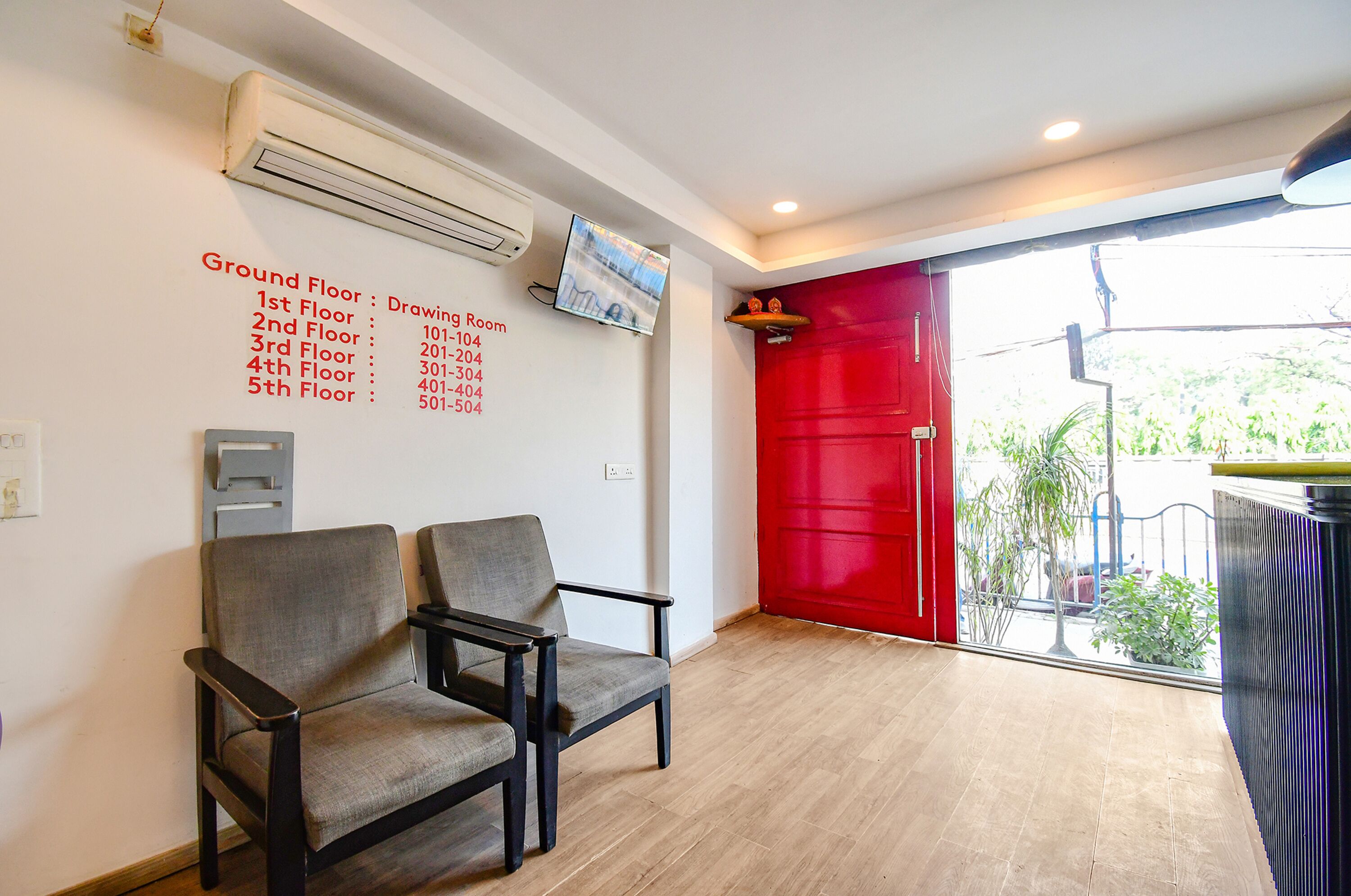 Un área de recepción con una puerta roja, dos sillas grises, un televisor montado en la pared y un cartel que muestra los números de habitación por piso.
