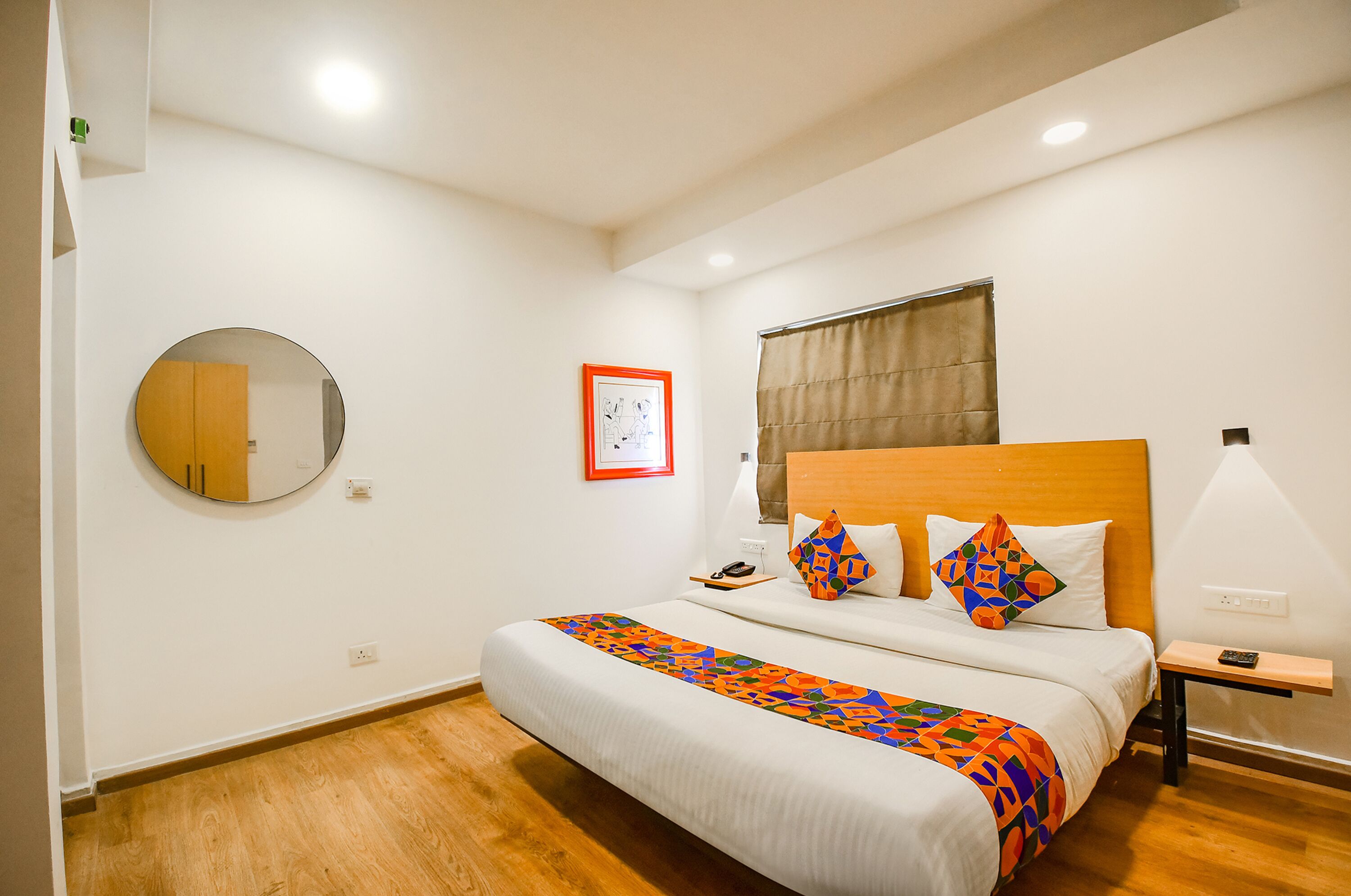 A bedroom with a wooden headboard, a round mirror, a bed with colorful bedding, a bedside table with a lamp, and a window with curtains.