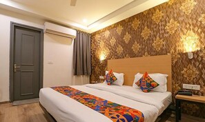 A hotel room with a bed, a nightstand, a lamp, and a door. - Fabexpress La Serene (New Delhi)