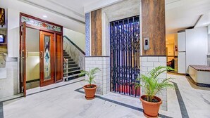 Un vestíbulo de hotel con piso de mármol, puerta de madera con paneles de vidrio, reja metálica y dos plantas en macetas.