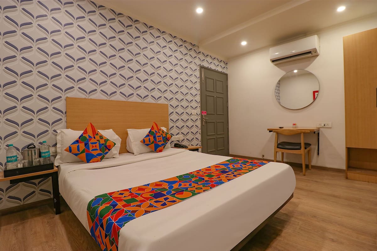 Habitación de hotel con una cama grande, una mesita de noche con lámpara, un pequeño escritorio con silla, un equipo de aire acondicionado y un espejo en la pared.