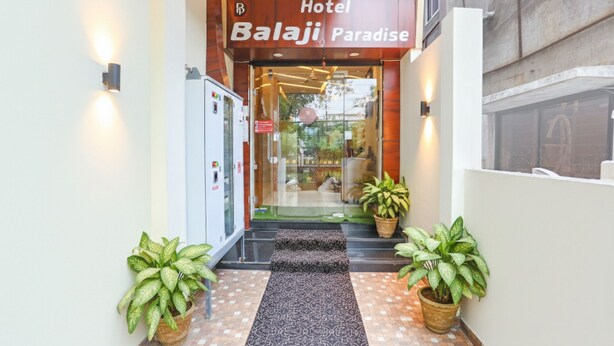 La entrada del Hotel Balaji Paradise, con un diseño moderno, presenta una puerta de vidrio, fachada de madera y un pequeño espacio exterior con plantas en macetas.