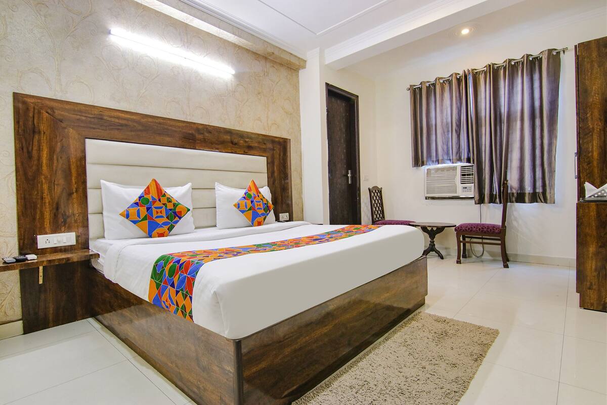 Une chambre d’hôtel avec un grand lit, une tête de lit en bois, une table de chevet, une chaise, une petite table et une fenêtre avec des rideaux.