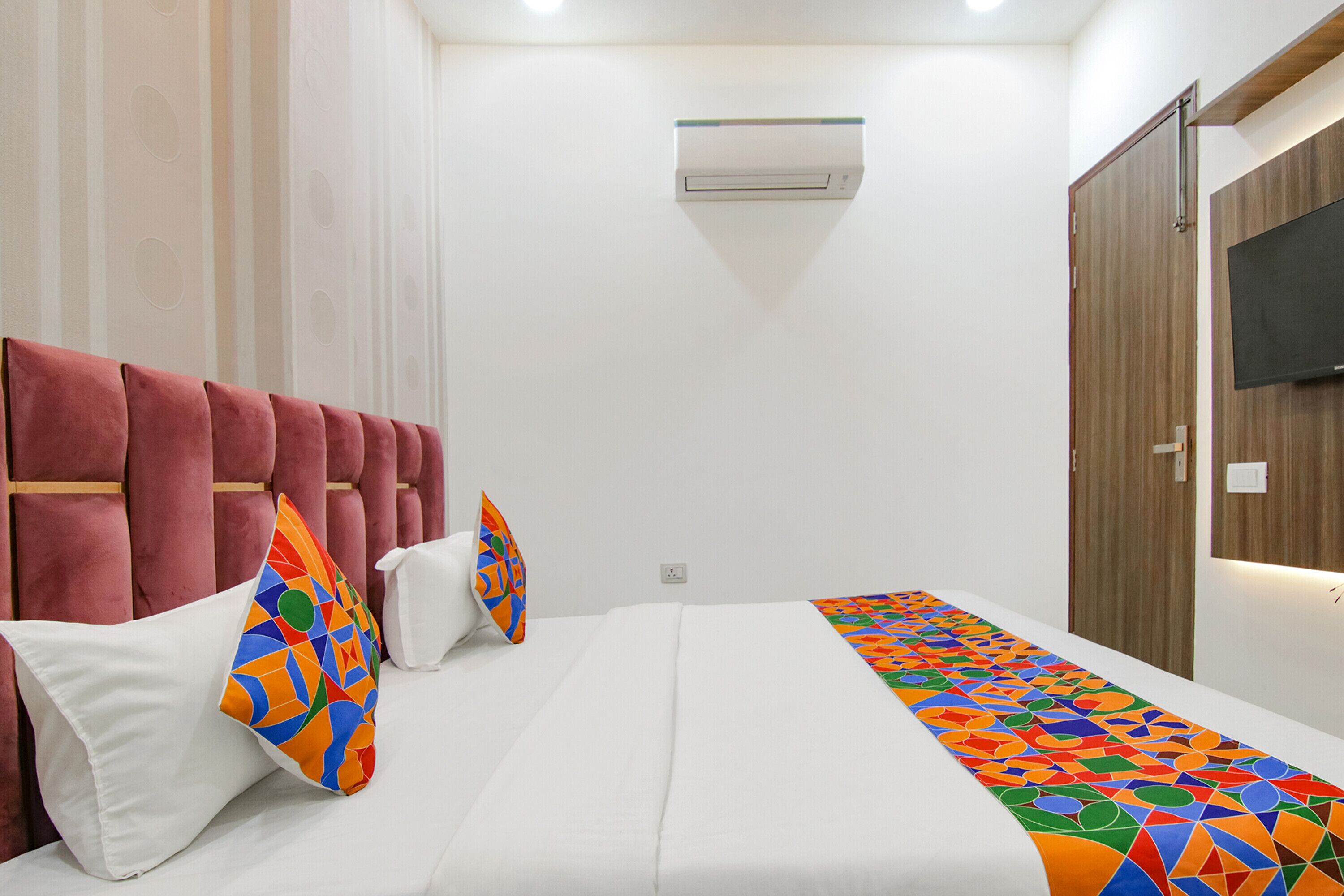 A hotel room with a bed, a red upholstered headboard, a white wall, and a mounted television.