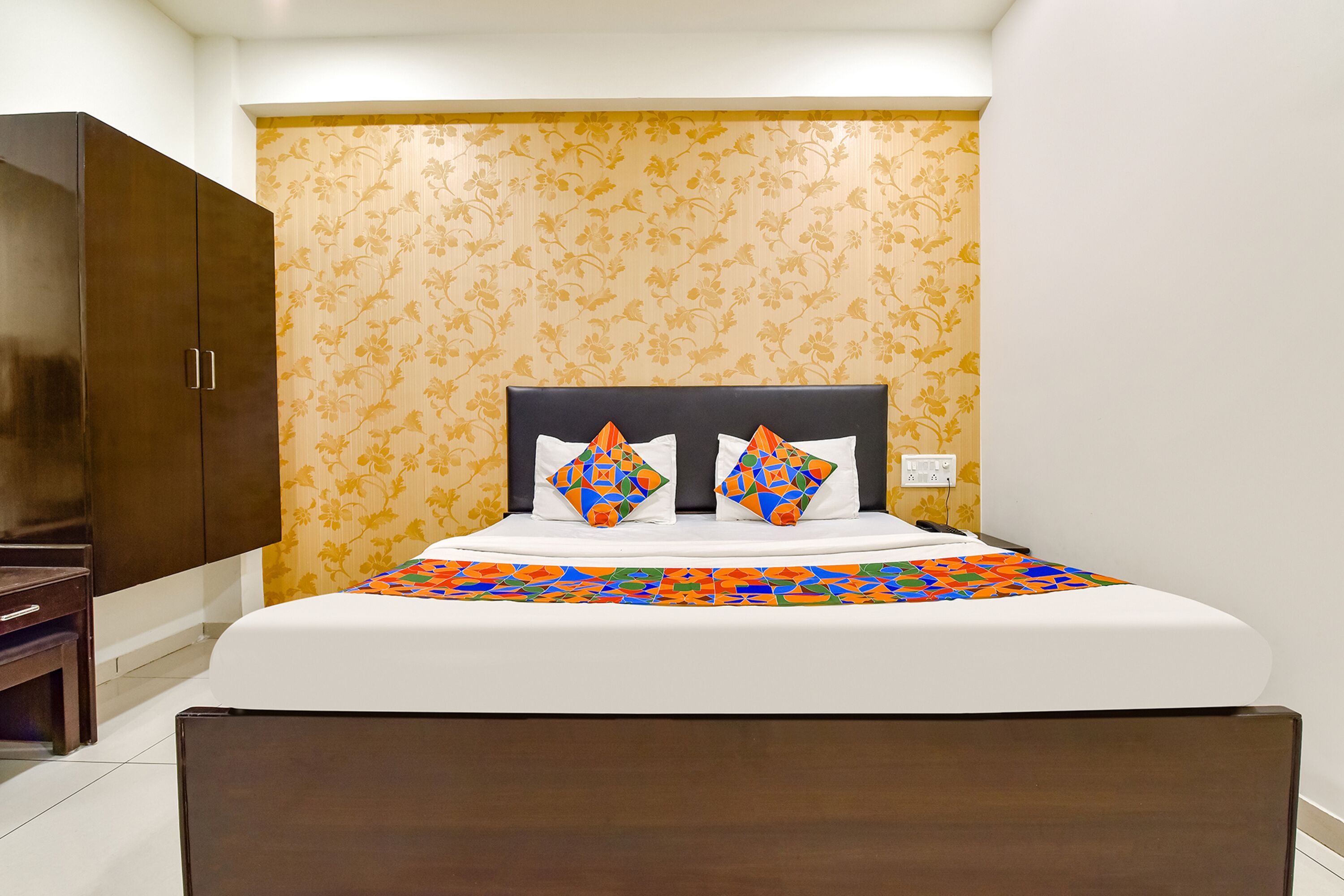 A neatly made bed with a patterned bedspread, a headboard with decorative pillows, and a wardrobe to the left.
