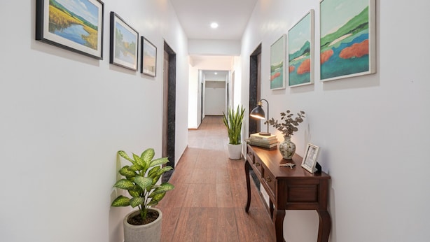 A hallway with framed landscape paintings on the walls, a potted plant, and a wooden side table with a lamp and books. - FabHotel Red Velvet & Suites (Pune)