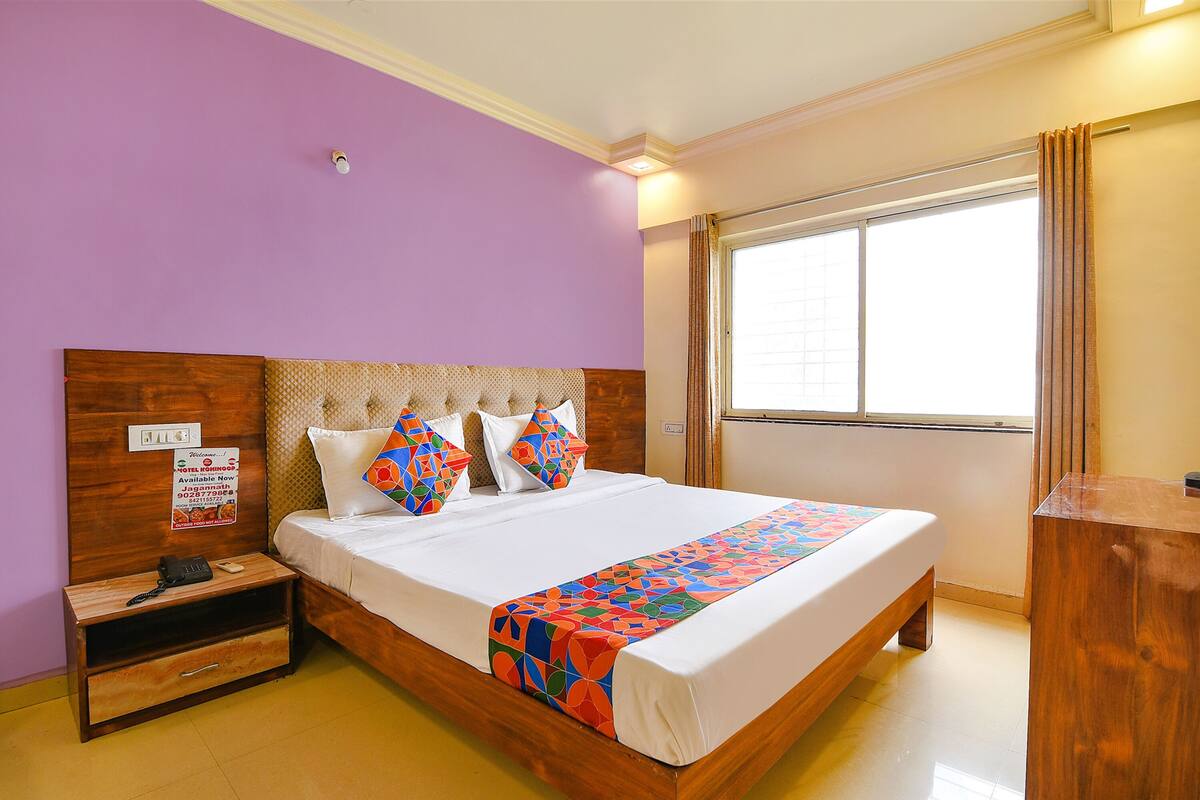 Quarto de hotel com cama, mesa de cabeceira, cabeceira de madeira, janela com cortinas e luminária fixada na parede.