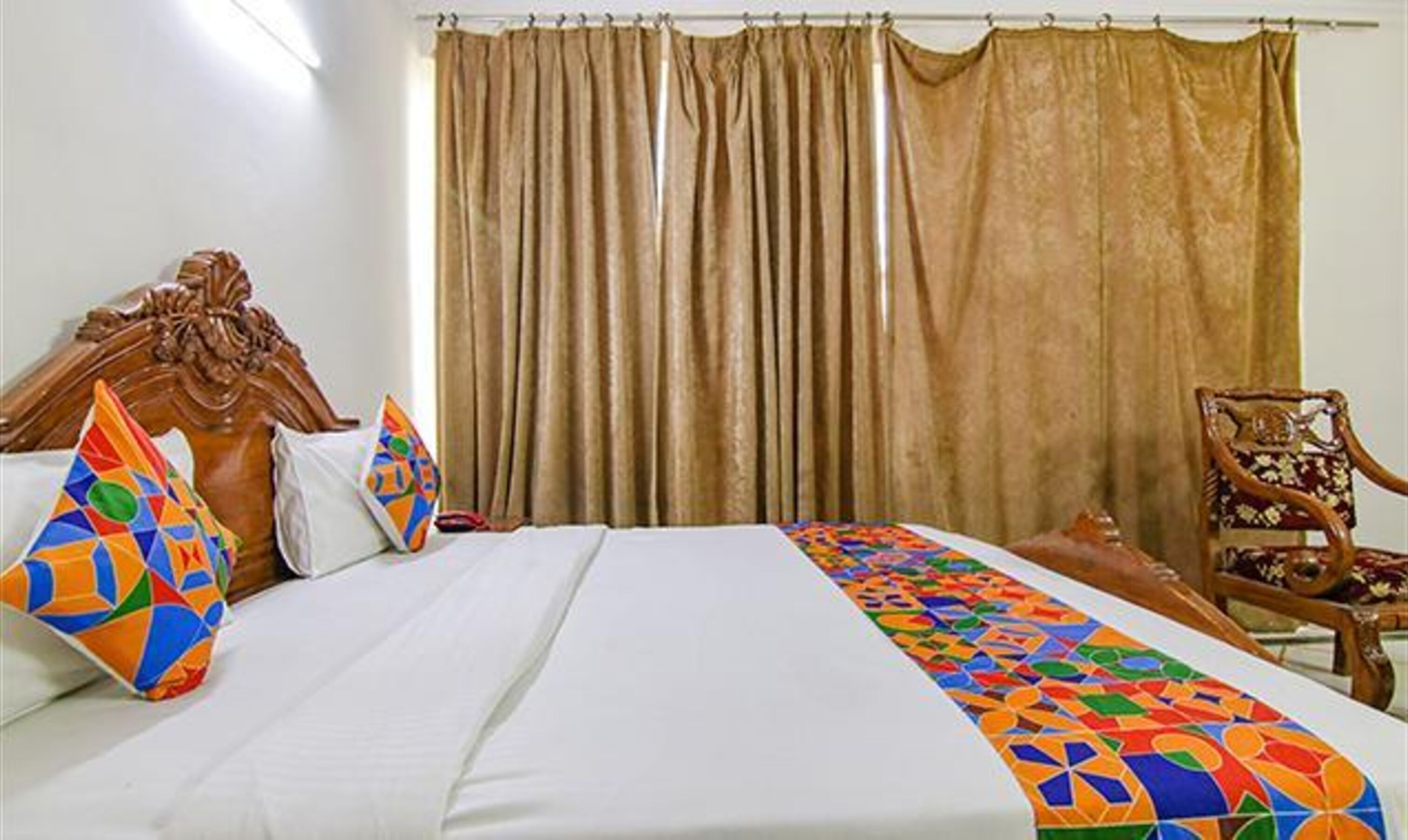 A hotel room with a bed, a wooden headboard, a colorful throw blanket, a wooden chair, and a large window with curtains.