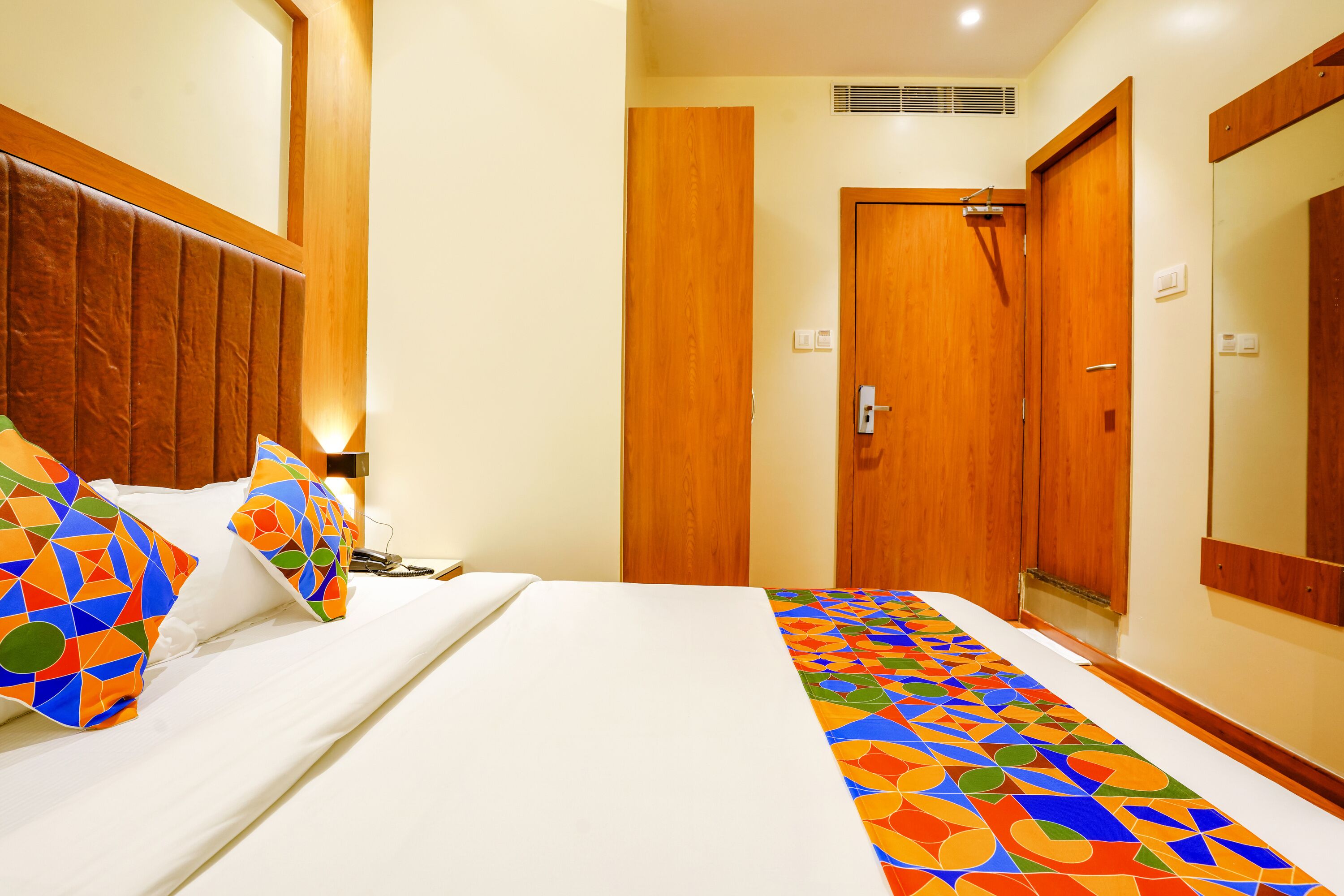 A hotel room with a bed, a wooden headboard, a door, and a mirror.