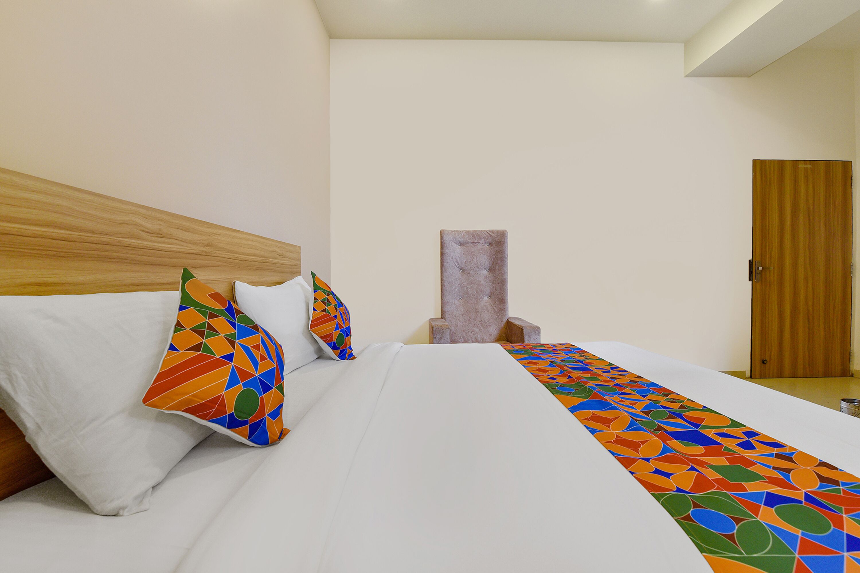 Un lit bien fait, avec du linge de lit blanc et des coussins aux motifs géométriques colorés, une tête de lit en bois et un mur sobre agrémenté d’une porte en bois.