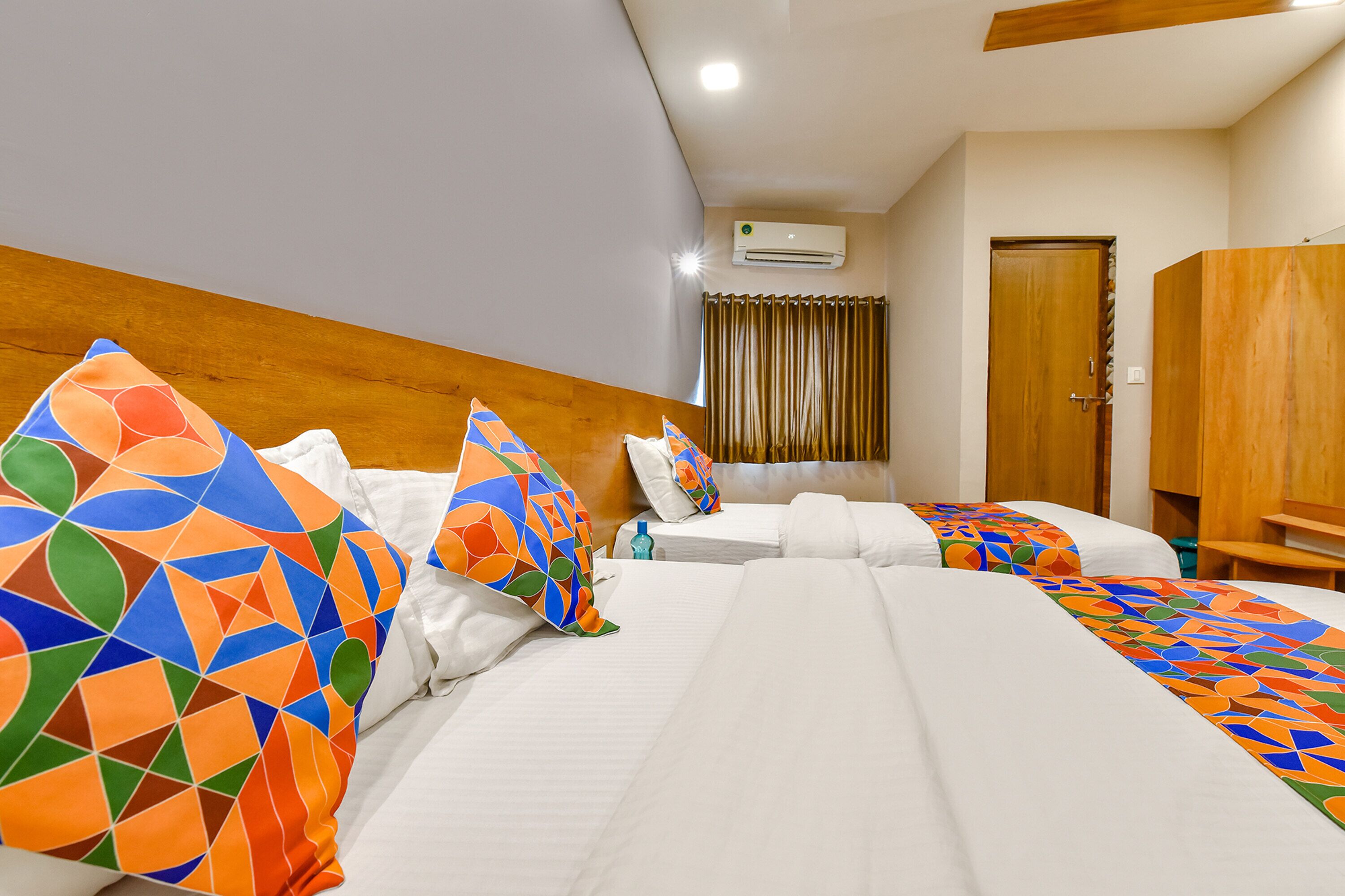 A hotel room with two beds, each with colorful geometric-patterned pillows, a wooden headboard, and a white bedspread.