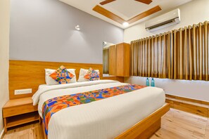 A modern hotel room with a wooden headboard, a large bed with colorful bedding, a built-in bench at the foot of the bed, a wooden floor, and a window with curtains. - FabHotel Gokul Villa (Ahmedabad)