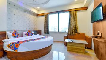 A modern hotel room with a curved bed, a wooden headboard, a flat-screen TV mounted on the wall, and a large window with curtains.