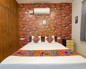 A bedroom with a brick-patterned wall, a bed with colorful pillows, a wooden wardrobe, and a small bedside table. - Fabexpress Kings Suites Hrbr Layout (Bengaluru)