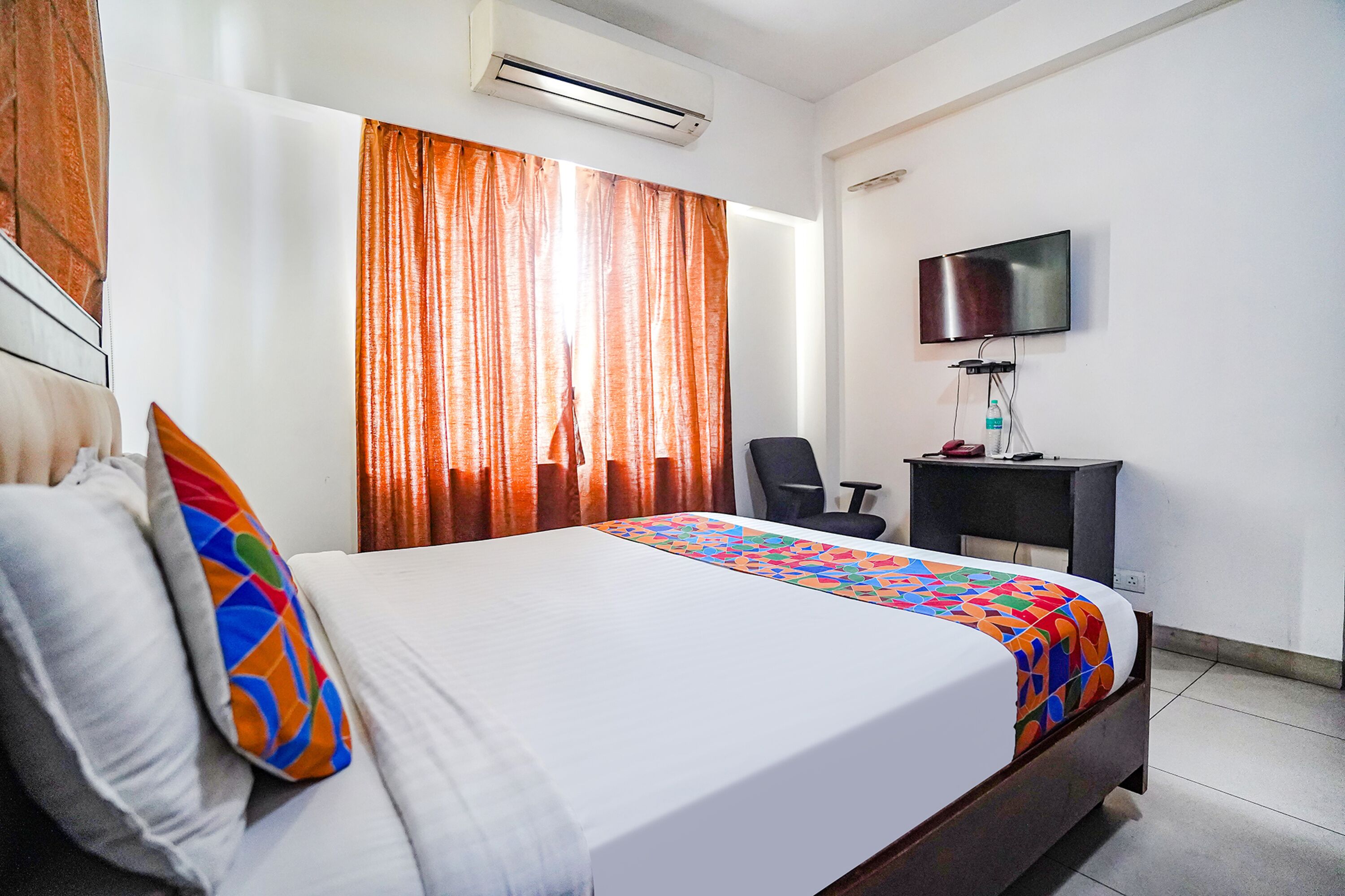 A hotel room with a bed, a desk, a chair, a TV, and an air conditioning unit.