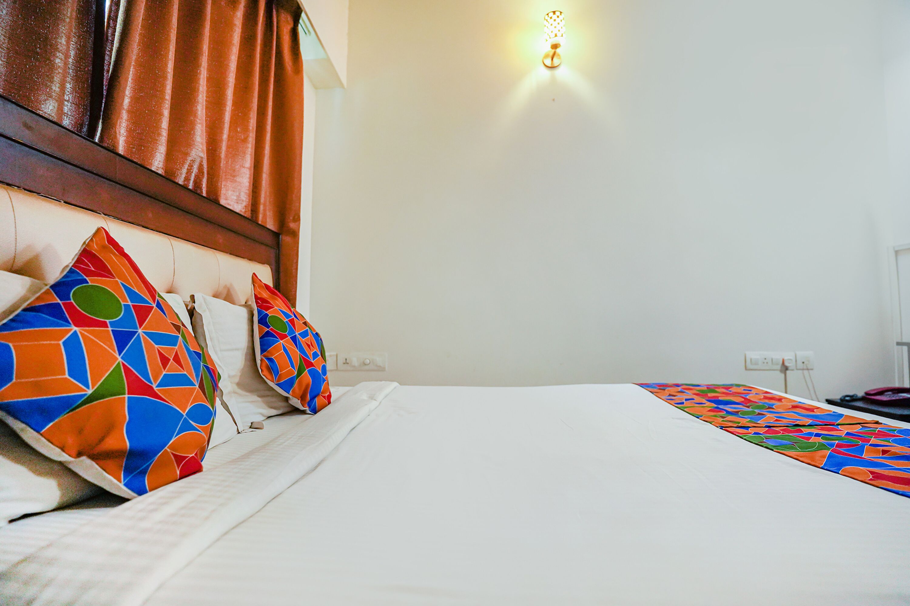 Une chambre d’hôtel avec un lit bien fait, des coussins colorés, un luminaire fixé au mur et un mur blanc uni.