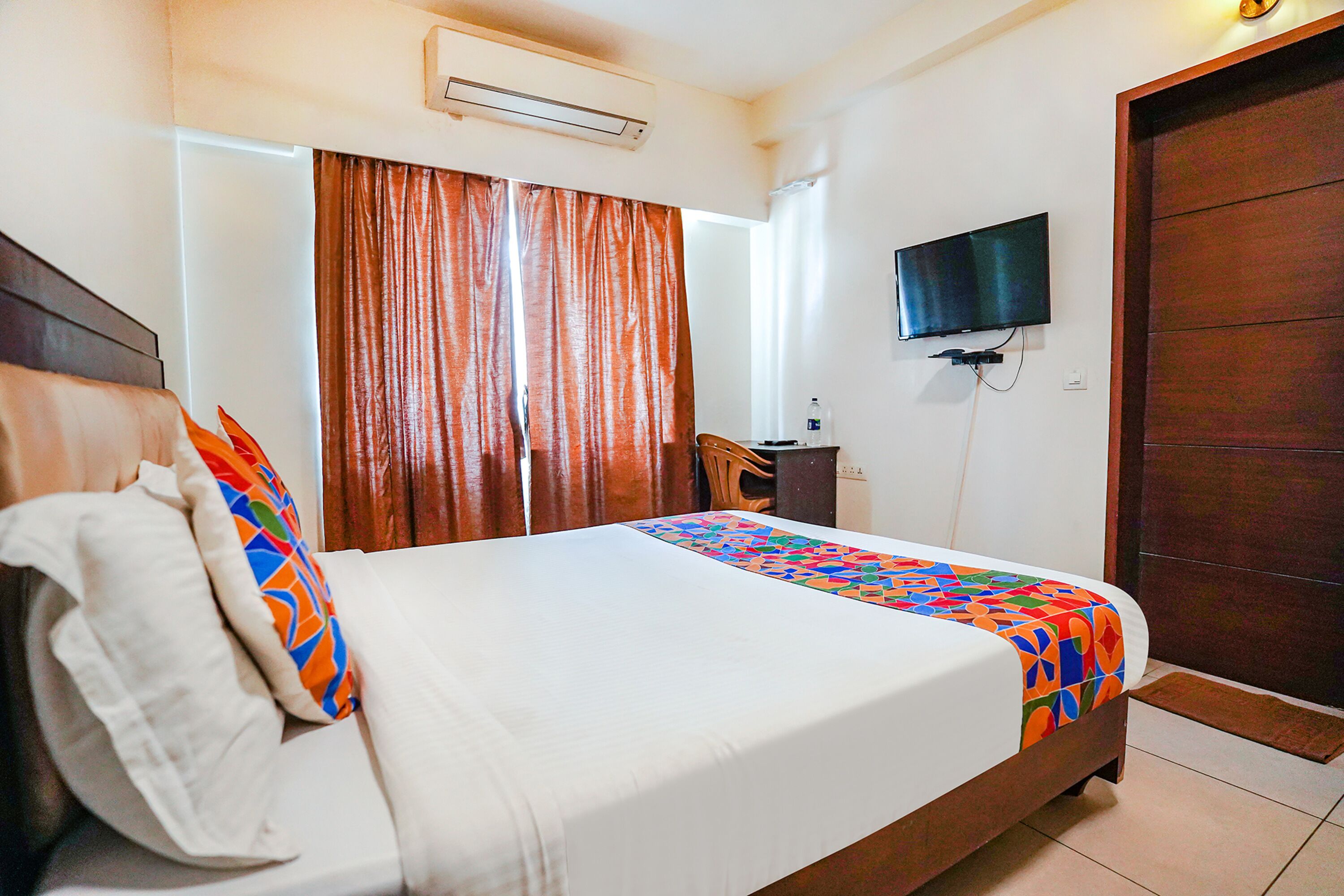 A neatly made bed with a colorful bedspread, a television mounted on the wall, and a wooden door.