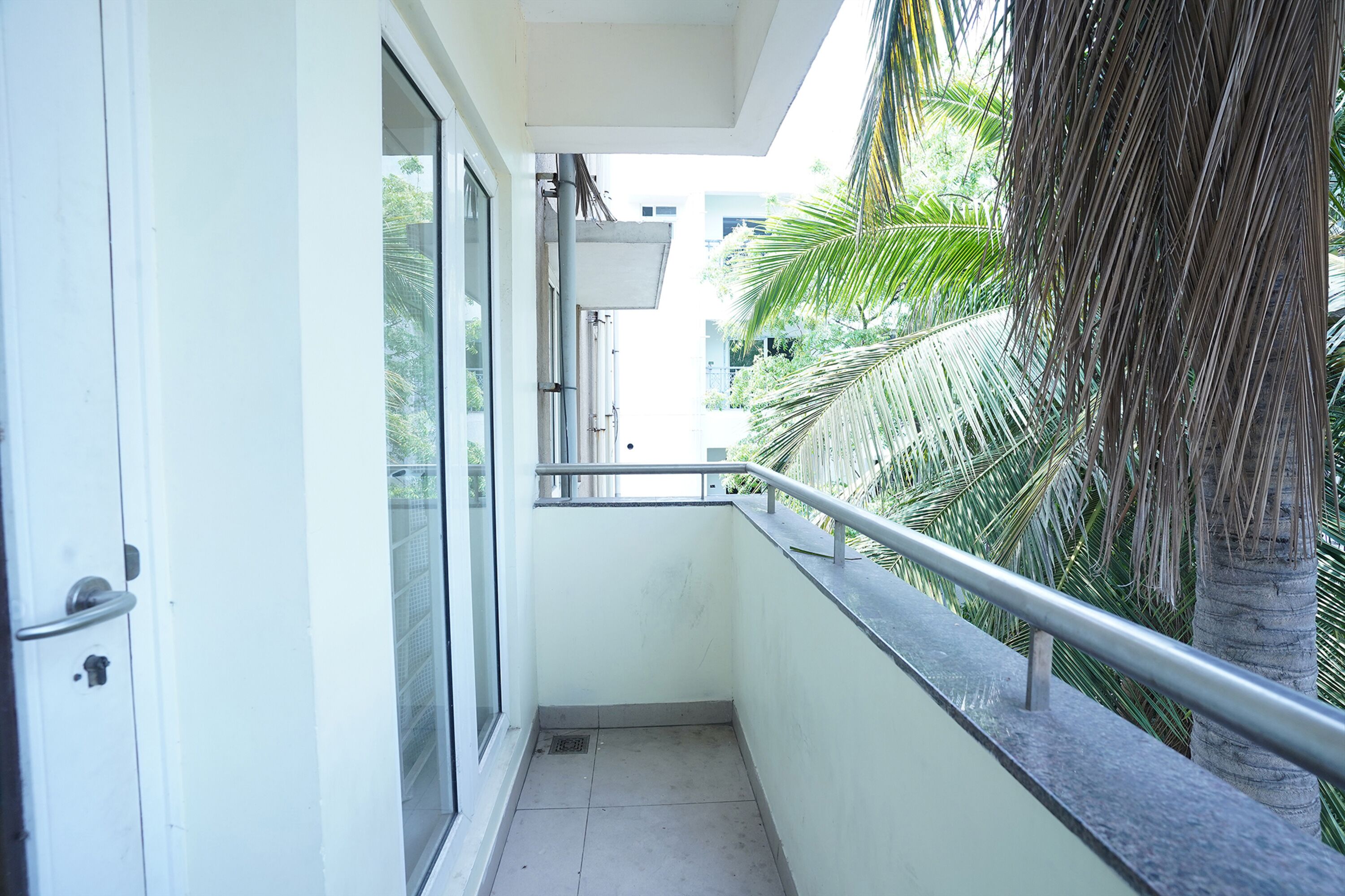 A narrow balcony with a metal railing, a glass door, and a view of palm trees and buildings in the background.
