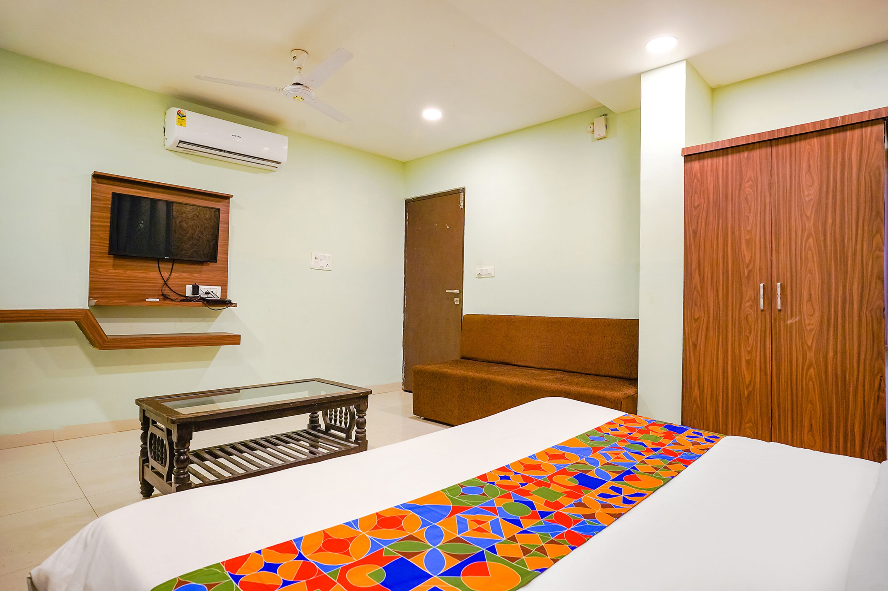 A bedroom with a bed, a wooden wardrobe, a brown sofa, a small table, and a mounted TV.