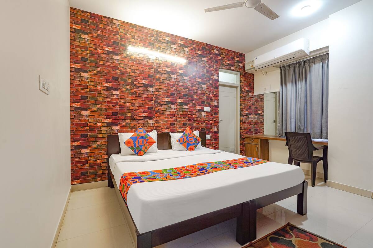 A room with a brick-patterned wall, a bed with a colorful bedspread, a desk with a chair, and a window with curtains.