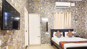A bedroom with a stone-patterned wall, a flat-screen TV mounted above the bed, a bedside table with a remote control, and a bed with colorful pillows. - FabHotel Kings Suites Banaswadi (Bengaluru)