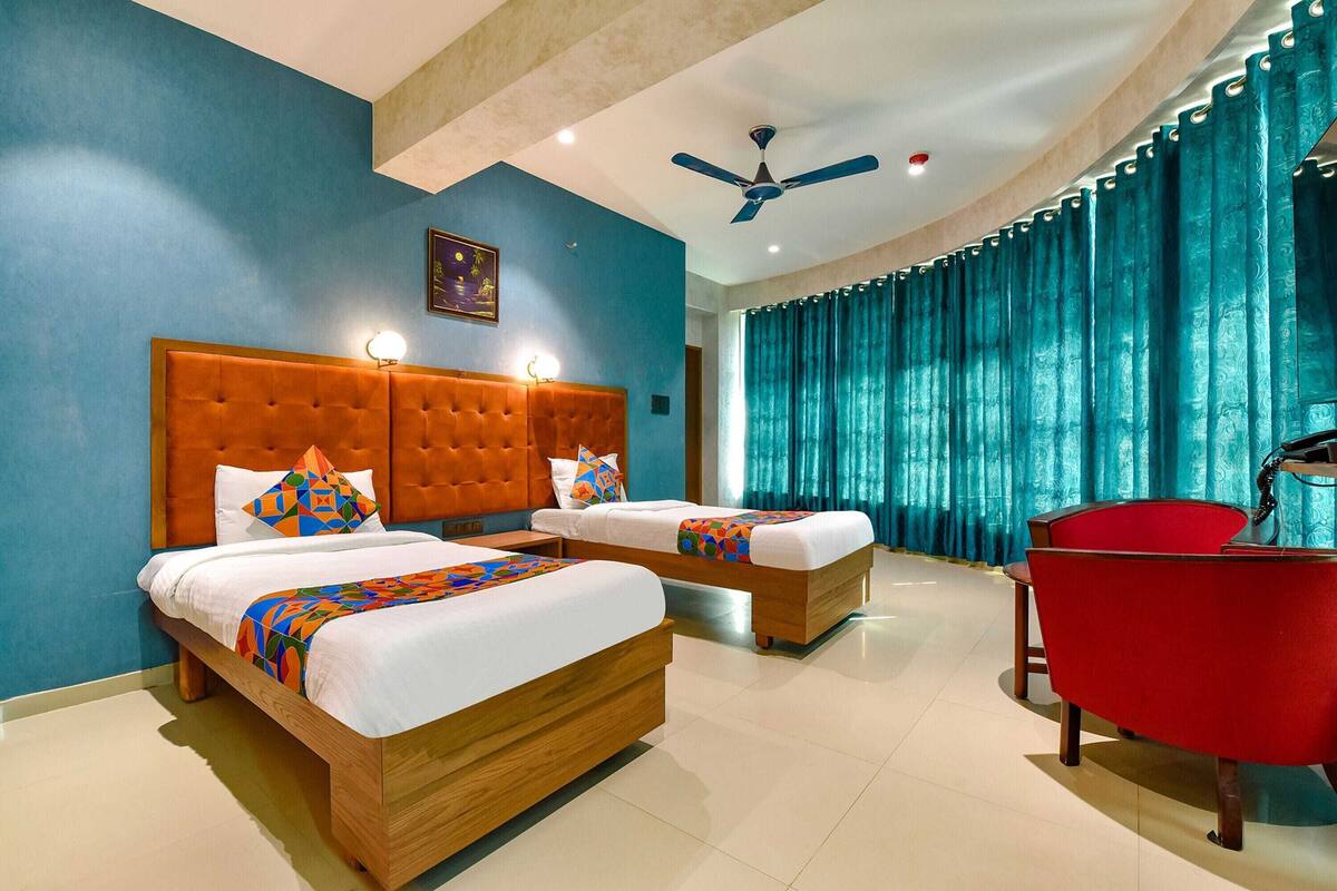 A hotel room with two beds, a red chair, a ceiling fan, and teal curtains.