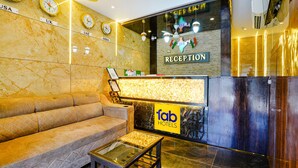 Zona de recepción de un hotel con un sofá, una mesa de centro de cristal, un mostrador de recepción y relojes que muestran diferentes husos horarios.