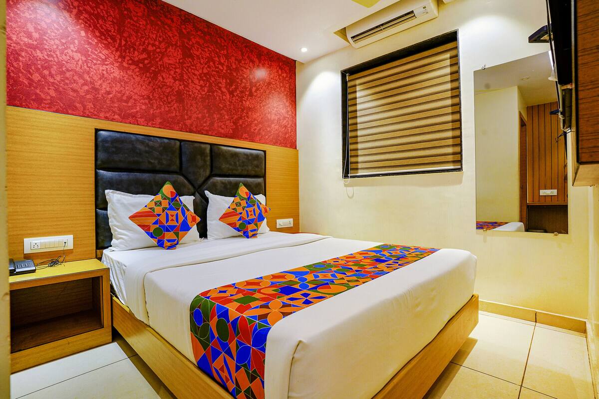 Habitación de hotel con una cama grande, cabecera de madera, pared de textura roja, ventana con persianas y una mesita de noche con teléfono.