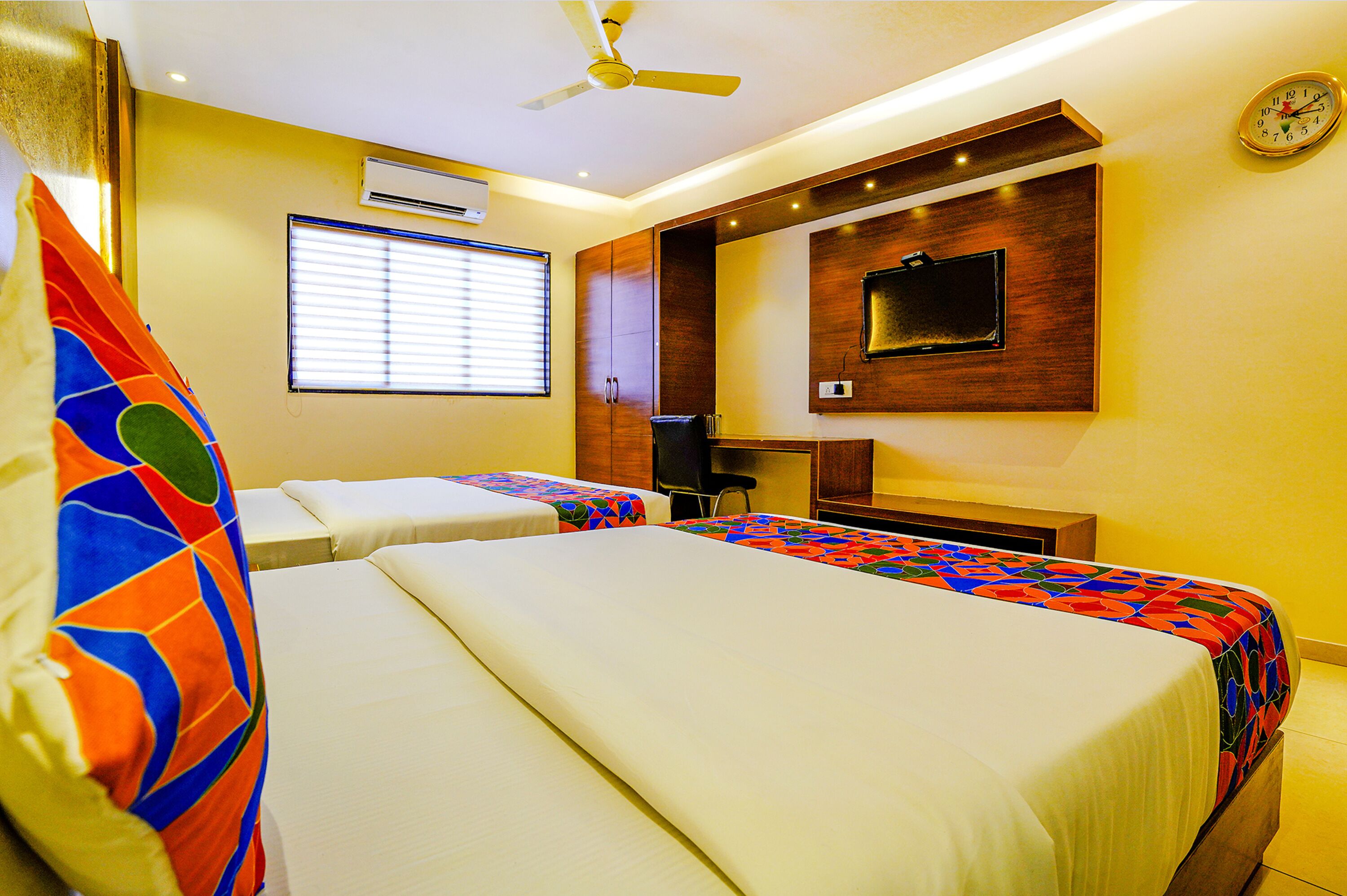 A hotel room with two beds, a ceiling fan, an air conditioning unit, a desk, and a television mounted on the wall.
