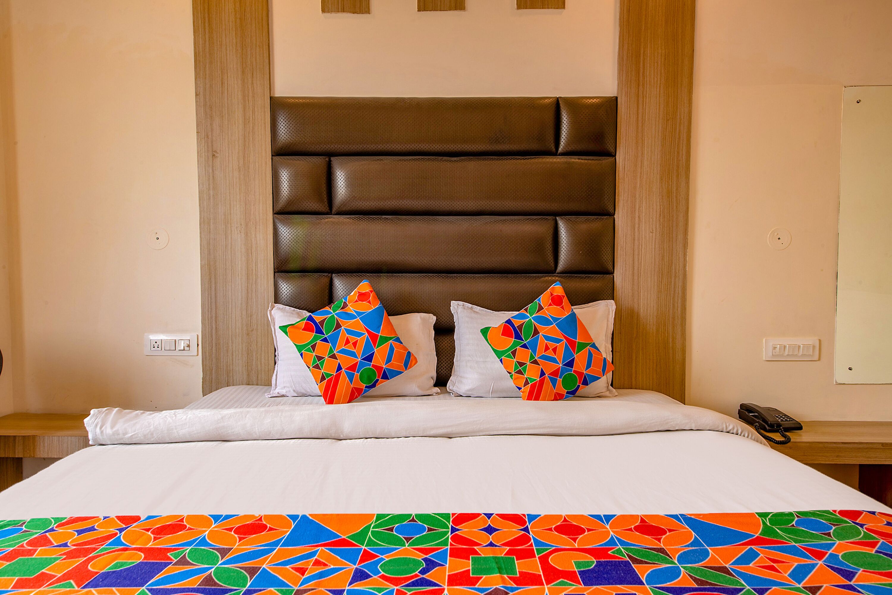 A hotel room with a large bed, a headboard, two pillows with colorful patterns, a bedside table with a phone, and a wall socket.