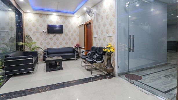 Un área de recepción con un sofá negro, un sillón negro, una mesita, un jarrón con flores, un televisor montado en la pared y una puerta de cristal que da a una zona de ducha.