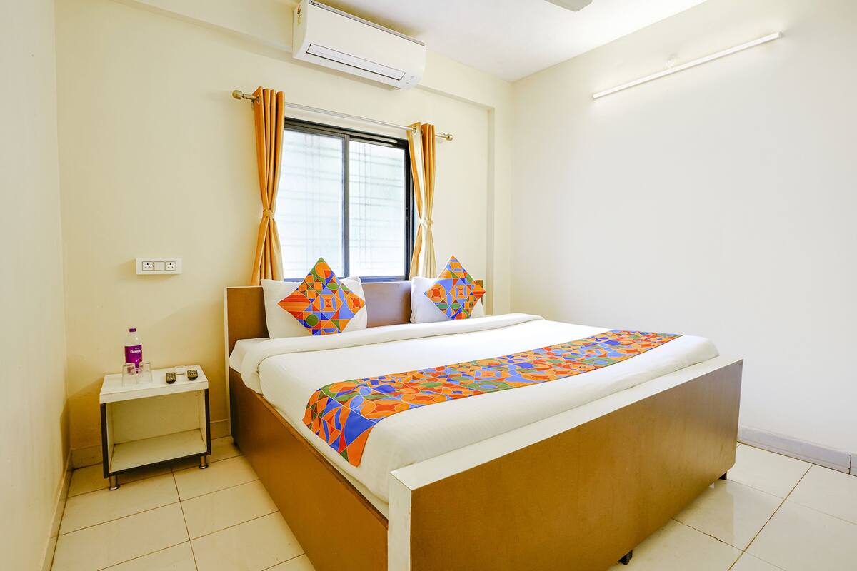 A neatly made bed with a colorful geometric-patterned bedspread, a bedside table with a bottle and remote control, and a window with curtains.