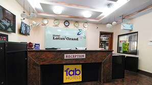 A hotel reception desk with a sign for 'Hotel Lotus Grand' and 'Fab Hotels'. There are clocks on the wall, a TV, and a digital clock displaying the time. - Fabhotel L Grand (Hyderabad)