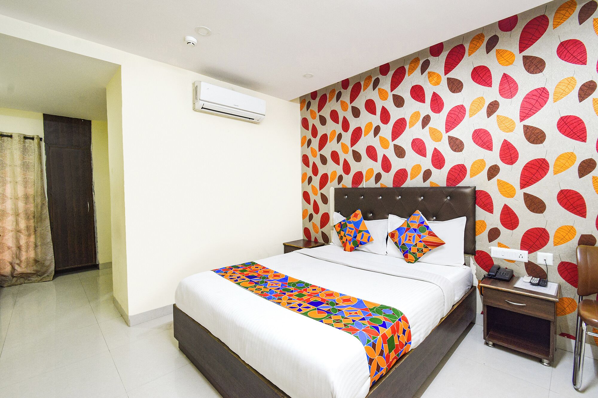 A hotel room with a bed, a nightstand, an air conditioning unit, and a wall with a leaf pattern.