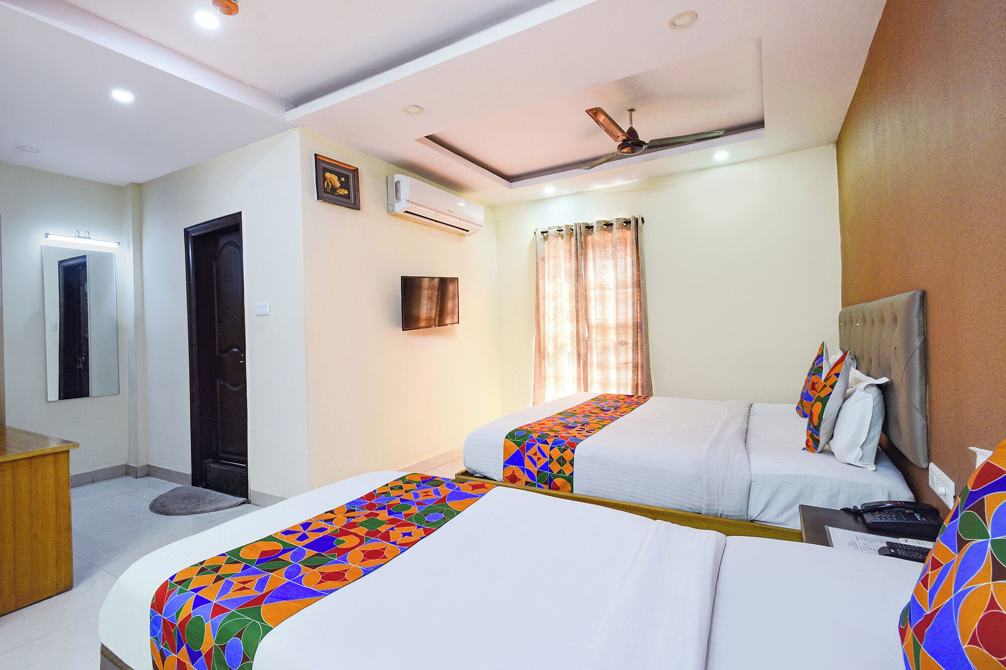 Une chambre d’hôtel avec deux lits, un bureau, un miroir, un climatiseur et un ventilateur de plafond.