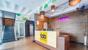 Mostrador de recepción de hotel con acabado de madera y un letrero de 'fab hotels'. Una escalera a la izquierda y una pared con un reloj y un letrero de Wi-Fi.