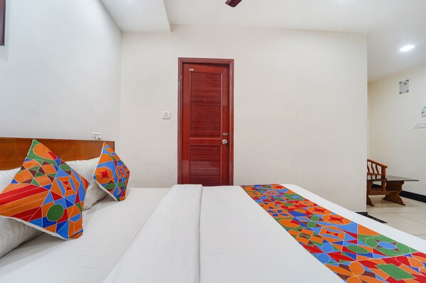 Una habitación de hotel con una cama, almohadas de diseño geométrico y colores vivos, un cabecero de madera, una puerta roja y una pequeña mesa con una silla.