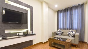 A modern living room with a flat-screen TV mounted on a marble wall, a wooden coffee table, a grey sofa with patterned cushions, and a window with grey curtains. - FabHotel Prime AV Royale (Dehradun)